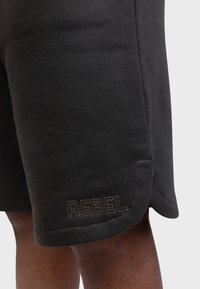 Shorts en coton noir avec une texture lisse. Dotés d'un logo brodé "REBEL" en marron foncé à l'ourlet. Finissions soignées pour les poignets et les poches.