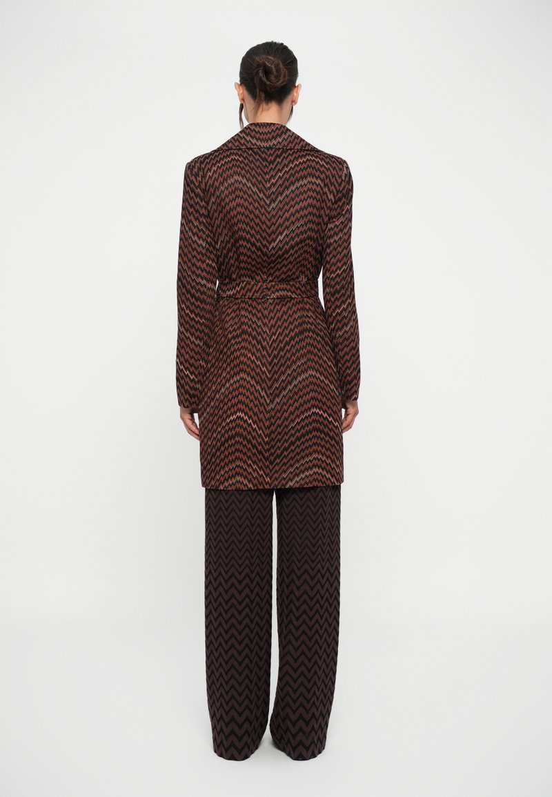 Femme avec les cheveux en chignon, portant un manteau à motifs zigzag marron et noir avec ceinture et un pantalon large assorti, se tenant debout devant un fond blanc uni.