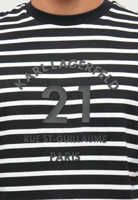 Juodai baltas horizontalių juostelių marškinėliai su iškilniu tekstu "KARL LAGERFELD 21 RUE ST-GUILLAUME PARIS" ant krūtinės.