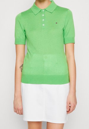 Personne portant un polo vert vif à manches courtes et une jupe blanche, debout devant un fond clair uni.