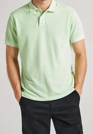 Homme portant un polo vert clair à manches courtes avec col et un pantalon noir, les mains dans les poches, debout devant un fond uni.