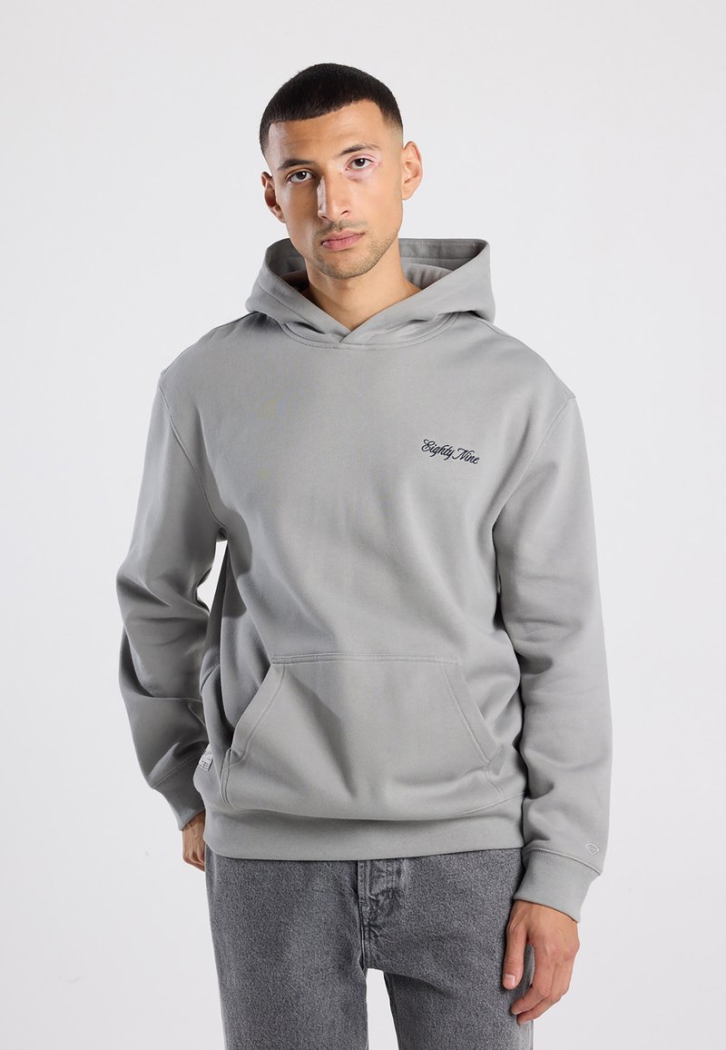 Uomo che indossa un felpa con cappuccio grigio chiaro con tasca frontale e testo in nero sul petto, abbinata a jeans grigio scuro, in piedi contro uno sfondo bianco.