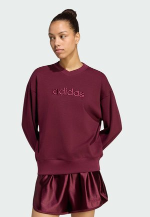 Kvinde iført en burgundy Adidas sweatshirt og matchende skinnende shorts, stående med hænderne bag ryggen mod en ensartet lys baggrund.