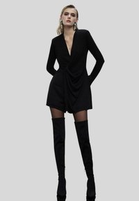 Jumpsuit nero a maniche lunghe con scollatura a V profonda, frontale drappeggiato e pantaloni shorts aderenti, abbinato a stivali neri alto fino alla coscia e grandi orecchini d'argento.