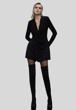 Jumpsuit nero a maniche lunghe con scollatura a V profonda, frontale drappeggiato e pantaloni shorts aderenti, abbinato a stivali neri alto fino alla coscia e grandi orecchini d'argento.