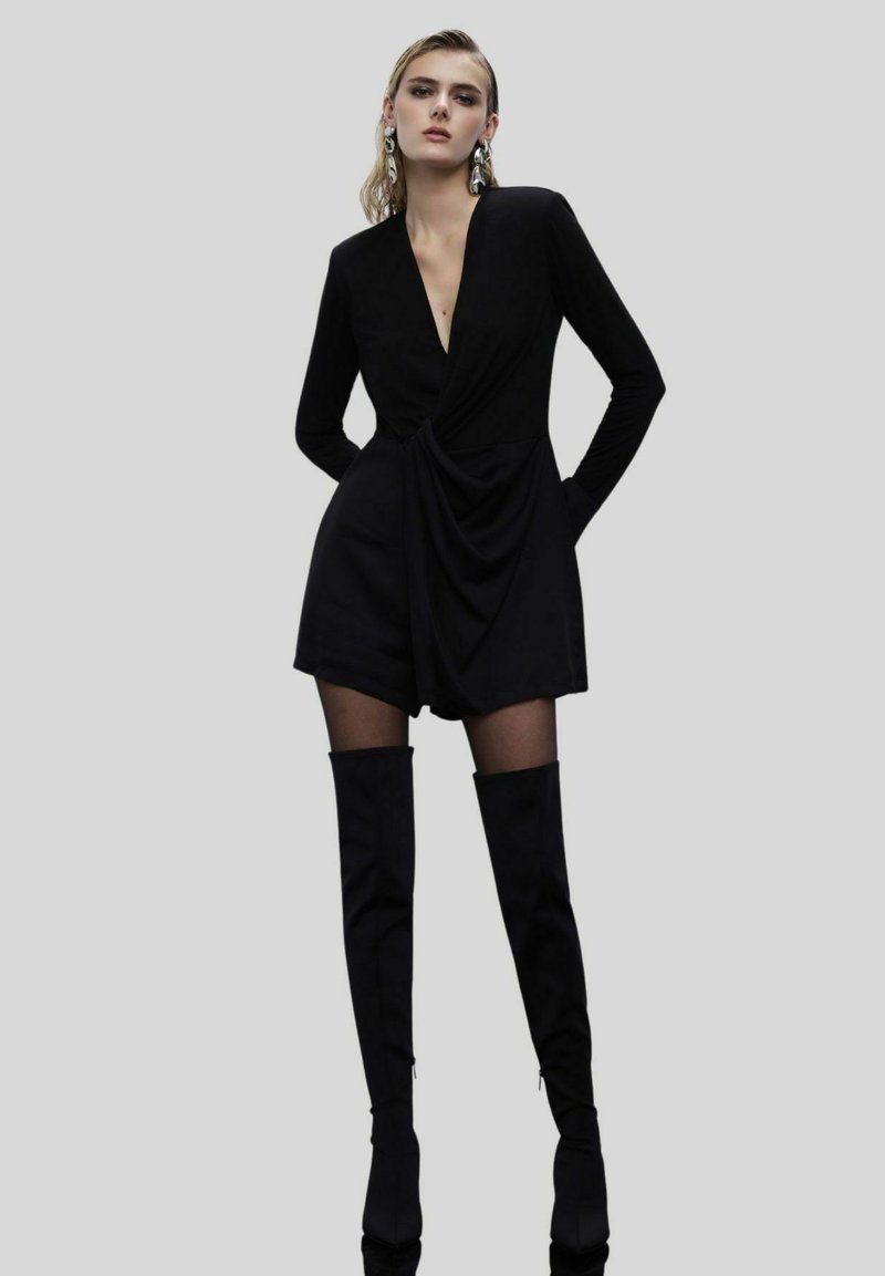 Jumpsuit nero a maniche lunghe con scollatura a V profonda, frontale drappeggiato e pantaloni shorts aderenti, abbinato a stivali neri alto fino alla coscia e grandi orecchini d'argento.