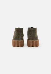 Grüne Wildleder-Ankle-Boots mit einer gerippten braunen Sohle, die einen hohen Kragen und minimale Nahtdetails auf der Rückseite aufweisen.