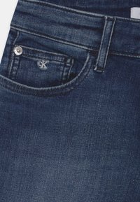 Tummansiniset farkut, joissa on etutasku hopeanvärisellä nappiq ja pieni CK-logo sen yläpuolella. Farkuissa on sileä pinta ja klassinen muotoilu.