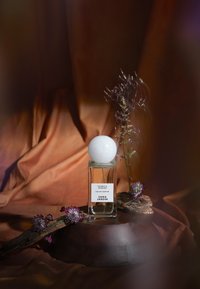 Szklana butelka perfum z białą kulistą zakrętką, z etykietą "VANILLA NOMAD" i "EAU DE PARFUM." Otoczona suszonymi kwiatami i drewnem.