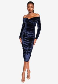 Jūras spēku zīda kleita ar plecus atsedzošu dekoltē, garām piedurknēm un sānu drapējumu. Iespējo pieguļoša silueta un garums līdz pusceļam. Caurspīdīgas siksnu kurpes.