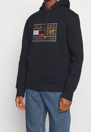Osoba nosząca czarną bluzę z kapturem z łatką z złotą obwódką, na której jest słowo "HILFIGER,", czerwona, biała i niebieska flaga oraz rzymskie cyfry MCMLXXV.