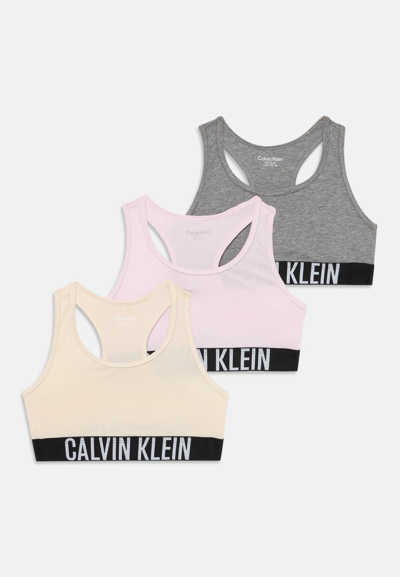 Tres sujetadores deportivos de Calvin Klein en gris, rosa claro y crema. Cada uno tiene una banda elástica negra con "CALVIN KLEIN" impresa en blanco. Diseño de espalda nadadora.