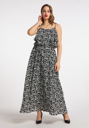 Vrouw draagt een zwarte maxi-jurk met witte bloemenprint, spaghettibandjes en ruches, gecombineerd met zwarte hoge hakken en gouden sieraden.