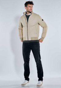 Beige Bomberjacke mit hohem Kragen, Reißverschluss vorne, elastischen Bündchen und Saum. Darüber getragen ein dunkelgrüner Pullover und schwarze Hose, kombiniert mit Sneakers.