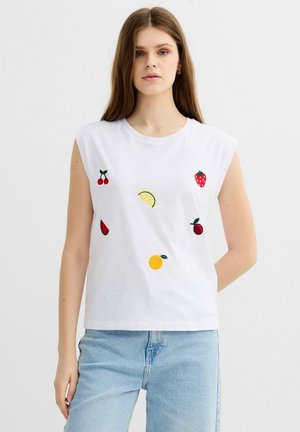 Joven mujer con top blanco sin mangas con diseños bordados de frutas y vaqueros azul claro sobre fondo liso.