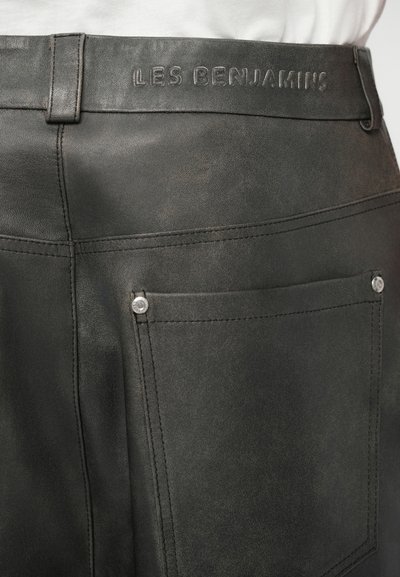 Pantalones de cuero negro con una textura suave, costuras en contraste, bolsillo trasero con botón a presión y logo de la marca en relieve en la cinturilla.