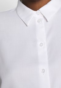 Weißes Hemd mit aufgestellten Kragen, aus glattem Stoff gefertigt. Verfügt über klare Kunststoffknöpfe und ein sauberes, schlichtes Design.