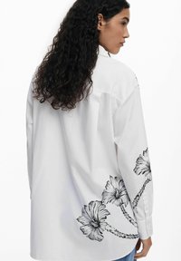 Camicia bianca a maniche lunghe con orlo curvo, caratterizzata da ricami floreali neri sul lato sinistro. Design con bottoni, realizzata in un tessuto leggero.