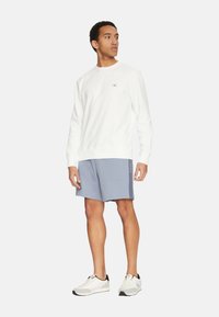 Sudadera blanca con cuello redondo y un pequeño logo en el pecho, combinada con pantalones cortos grises. El modelo está de pie con zapatillas deportivas, mostrando un diseño simple.