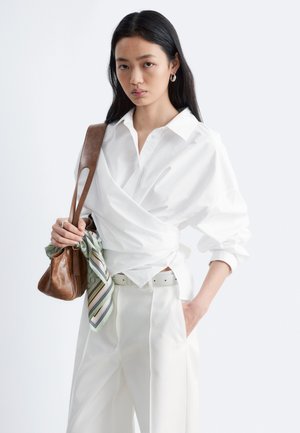 Kobieta o długich czarnych włosach w białej bluzce typu wrap i spodniach, niesie brązową skórzaną torebkę z paskiem w paski, ma rękę w kieszeni.