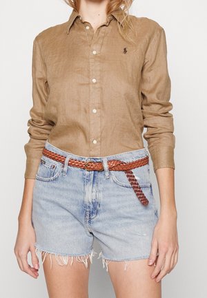 Femme portant une chemise boutonnée couleur sable rentrée dans un short en denim bleu clair avec des ourlets effilochés et une ceinture tressée marron.