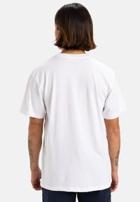 Camiseta de algodón blanca con mangas cortas, cuello redondo y un corte holgado. Muestra tatuajes en los brazos de la persona que la lleva puesta.