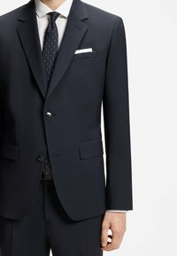 Homme portant un costume bleu marine foncé avec une chemise blanche, une cravate noire à motifs et un mouchoir blanc sur fond blanc uni.