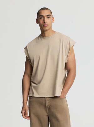 Giovane uomo con capelli corti che indossa una maglietta beige senza maniche e pantaloni marroni, in piedi con una mano in tasca su uno sfondo semplice.