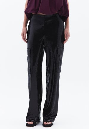 Pantalones cargo negros con lentejuelas y corte holgado, que presentan bolsillos laterales y una superficie brillante y texturizada. Combinados con una blusa morada y tacones negros.
