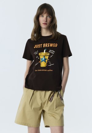 Kobieta o włosach do ramion nosi czarny T-shirt z grafiką filiżanki kawy i napisem „JUST BREWED”, zestawiony z beżowymi szortami.