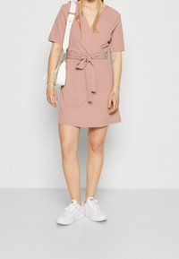 Krátké růžové zavinovací šaty s krátkými rukávy, páskem a hladkým materiálem, doplněné bílými teniskami a malou bílou taškou.