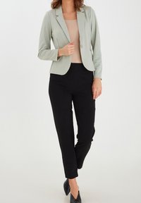 Blazer verde chiaro con colletto a rever, abbinato a pantaloni neri aderenti. Il tessuto appare liscio, con un design strutturato.
