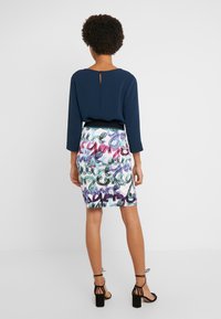 Blouse navy avec un dos en keyhole, associée à une jupe ajustée avec un imprimé abstrait multicolore. Des escarpins noirs à lanières complètent le look.