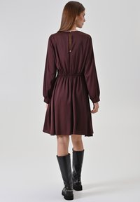 Dunkelburgunderrotes Kleid mit langen Ärmeln, tailliert und mit Rückenschlüssel-Lochausschnitt. Kombiniert mit knielangen schwarzen Stiefeln. Glatte Stofftextur.