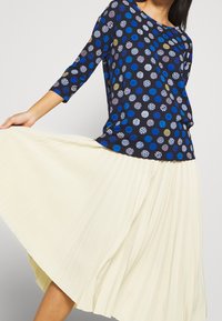 Marinebluse mit mehrfarbigen Punkten und 3/4-Ärmeln, kombiniert mit einem cremefarbenen Faltenrock. Das Outfit hat eine lockere Passform und eine glatte Textur.