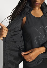 Personne ajustant le cordon intérieur d'une veste noire, portant un débardeur noir avec un motif de montagne et de mousqueton.