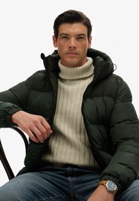 Puff jacket i mørkegrøn med hætte, båret over en struktureret cremefarvet rullekravesweater; modellen har også blå jeans og et ur på.