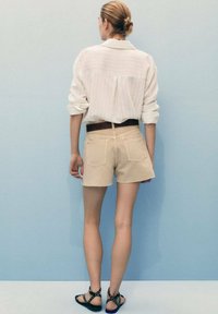 Chemise rayée blanche, shorts beiges, ceinture marron, sandales noires à clous, vues de dos sur fond bleu clair.