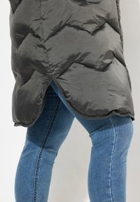 Schwarze Steppjacke mit gestepptem Muster und hohem Kragen, kombiniert mit blauen Jeans. Die Jacke hat einen leicht geschwungenen Saum am unteren Ende.