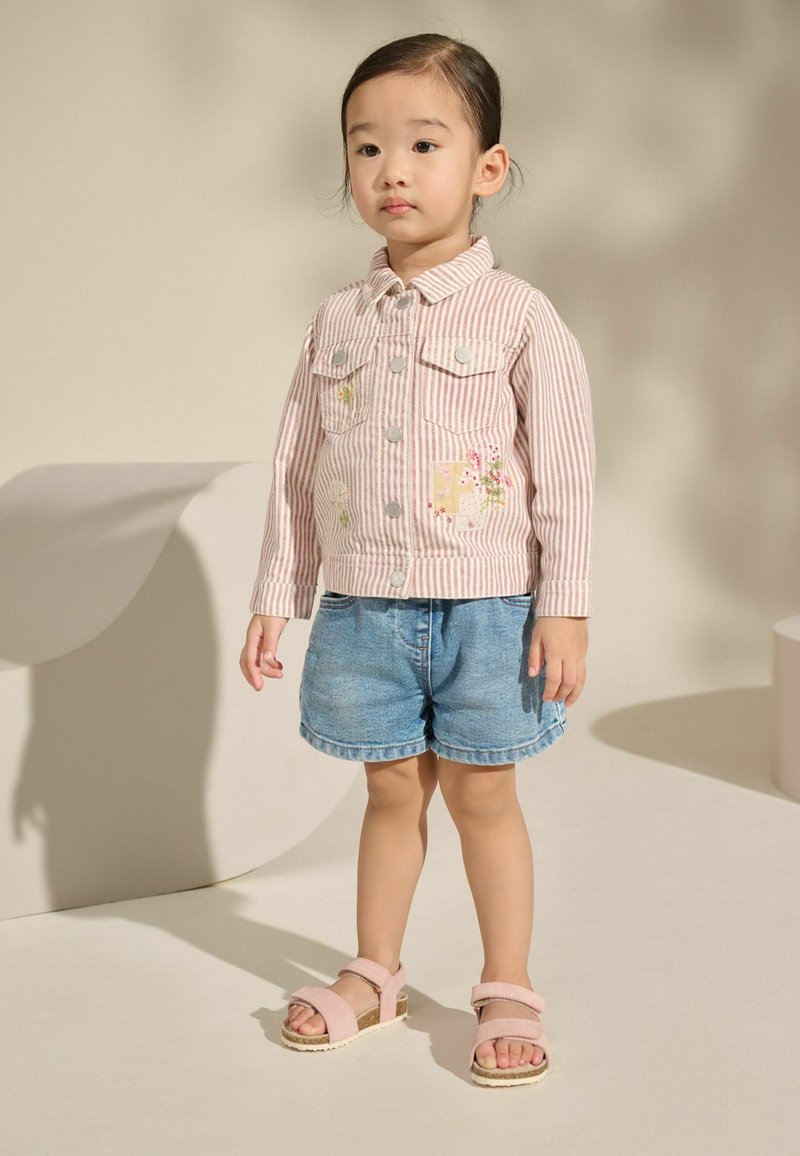Ein junges Kind steht, trägt eine rosa gestreifte Jacke mit Blumenapplikationen, Jeansshorts und rosa Sandalen vor einem schlichten beigen Hintergrund.