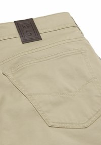 Beige Hose mit verstärkter Naht an der Gesäßtasche und einem braunen Lederetikett mit der Aufschrift "M5" auf dem Bund.