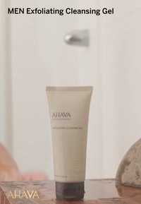 Gel esfoliante per la pulizia AHAVA MEN in tubo a compressione, color beige traslucido con cappuccio grigio scuro, presenta gocce d'acqua sulla superficie.