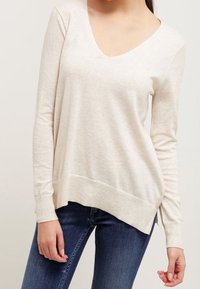 Pull beige clair à manches longues avec un col en V profond et un ourlet droit, présentant une coupe décontractée et un tissu en maille doux et texturé.