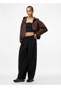 Felpa corta con zip marrone, top nero senza maniche e pantaloni larghi neri con pinces. Sneakers nere con design testurizzato.