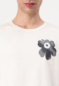 Ljusbeige t-shirt med rund halsringning, med en stor svart blommig grafik på vänster bröstområde. Mjuk, slät textur.
