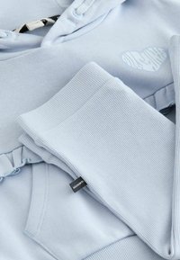 Hanorac albastru deschis cu manșete striate și un mic model cu inimă. Are glugă cu șnur și o etichetă inscripționată cu "ALLSAINTS". Textură moale.