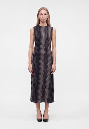Femme aux cheveux blonds lisses portant une robe midi sans manches à motif zigzag noir et gris et des talons noirs pointus, debout immobile.