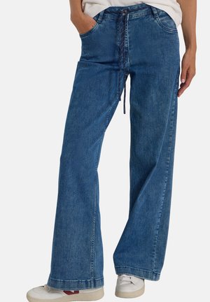 Personne portant un jean bleu à jambes larges avec une ceinture en corde et des baskets blanches, debout, une main dans la poche, sur un fond uni.