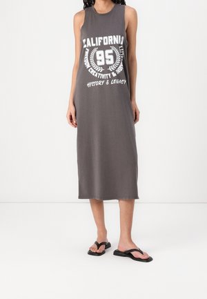 Frau trägt ein ärmelloses graues Midikleid mit „California 95“ und Textprint sowie schwarze Flip-Flops und steht vor einem schlichten Hintergrund.