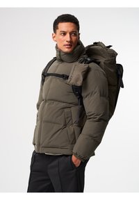 Olivgrüne Pufferjacke mit horizontalen Steppdesign, Reißverschlusstaschen und elastischen Bündchen, kombiniert mit einem schwarzen Rucksack mit verstellbaren Trägern.